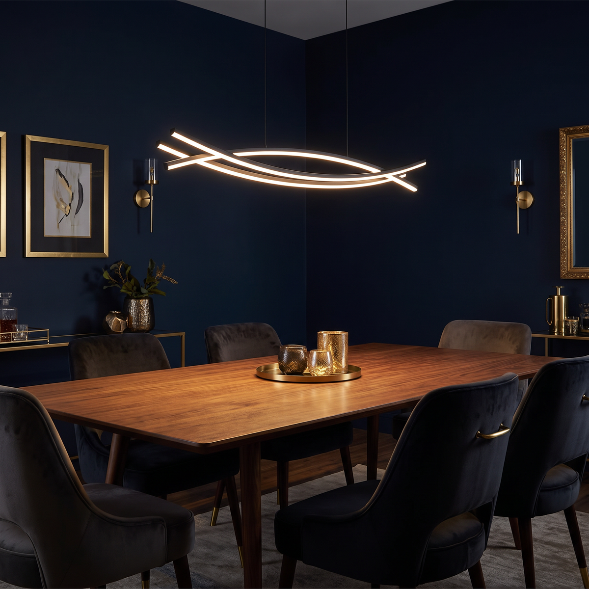 Marea wavy LED pendant light hangs low over a long walnut dining table in a dark moody dining room with deep navy walls