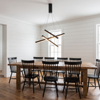 Criss crossed-bar linear LED chandelier hangs over a long dining table in a modern farmhouse dining room with white shiplap walls