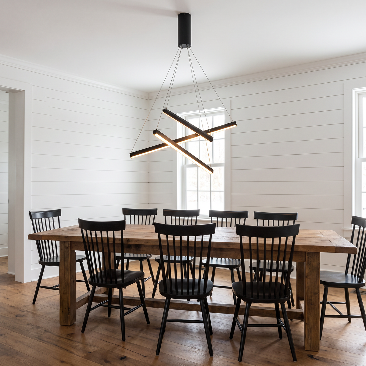 Criss crossed-bar linear LED chandelier hangs over a long dining table in a modern farmhouse dining room with white shiplap walls